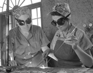 Two women in overalls, headscarves and big goggles solder together.