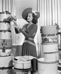 A woman in a 1940's dress and surrounded by hat boxes, chooses the one she wants.