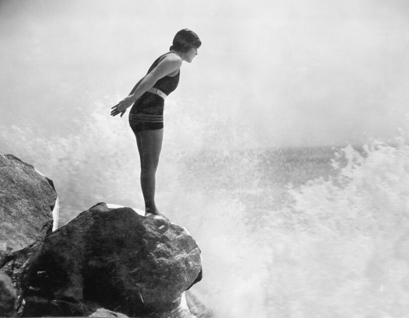 A woman in a swimming costume stands poised and ready to dive off a rock into the sea