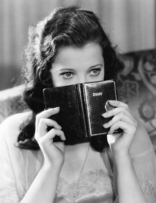 A dark haired woman from the 1940's peers over a small black book she is holding in front of her face.